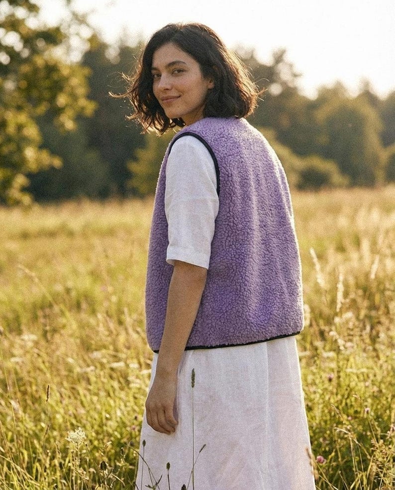 K&ouml;nnte beinhalten: Eine Frau tr&auml;gt eine lavendelfarbene Fleeceweste mit schwarzem Besatz &uuml;ber einem wei&szlig;en Kurzarmshirt und einem wei&szlig;en Rock. Sie steht in einem Feld mit hohem Gras, im Hintergrund B&auml;ume. Die Weste ist das zentrale Kleidungsst&uuml;ck.
