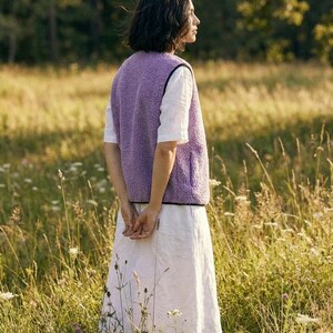K&ouml;nnte beinhalten: Eine Person tr&auml;gt eine lavendelfarbene Fleeceweste mit schwarzem Besatz &uuml;ber einem wei&szlig;en Kurzarmhemd und einem wei&szlig;en Rock. Die Person steht in einem Feld mit hohem Gras und Wildblumen.