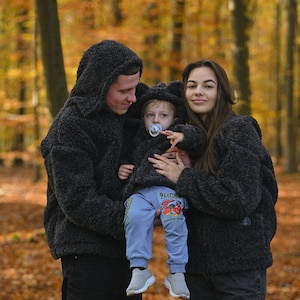 Puede incluir: Una familia de tres personas con chaquetas con capucha de color negro y pelusa a juego en un entorno de bosque. Los padres sostienen a un ni&ntilde;o peque&ntilde;o que lleva una chaqueta similar. El ni&ntilde;o lleva una camisa azul con un estampado gr&aacute;fico y un chupete.