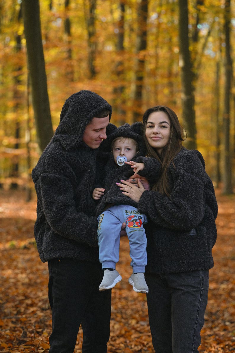 Puede incluir: Una familia de tres personas con chaquetas con capucha de color negro y pelusa a juego en un entorno de bosque. Los padres sostienen a un ni&ntilde;o peque&ntilde;o que lleva una chaqueta similar. El ni&ntilde;o lleva una camisa azul con un estampado gr&aacute;fico y un chupete.