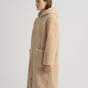 May include: A long, beige, hooded coat made of a textured, fleece-like material. The coat features a full-length front zipper closure, long sleeves, and two large, square patch pockets positioned on the front lower section of the garment.
