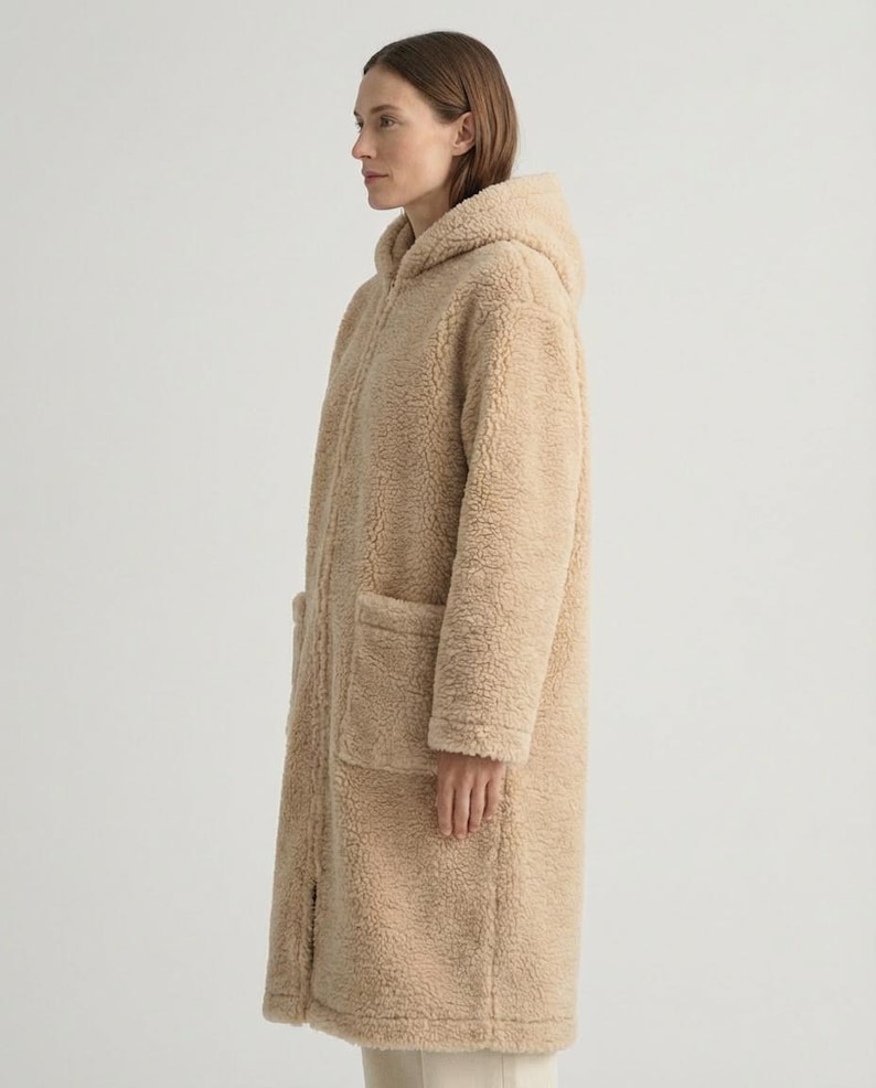 May include: A long, beige, hooded coat made of a textured, fleece-like material. The coat features a full-length front zipper closure, long sleeves, and two large, square patch pockets positioned on the front lower section of the garment.
