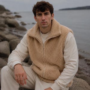 Könnte beinhalten: Eine ärmellose beigefarbene Sherpa-Fleece-Weste mit durchgehendem Frontreißverschluss und hohem Kragen, getragen über einem langärmeligen cremefarbenen Sweatshirt und einer passenden cremefarbenen Hose. Die Weste hat ein strukturiertes, flauschiges Material.
