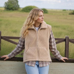 Könnte beinhalten: Eine ärmellose Weste aus beigem Sherpa-Fleece mit Stehkragen, durchgehendem Frontreißverschluss und zwei aufgesetzten Vordertaschen. Die Weste wird über einem langärmeligen, braun-weiß karierten Hemd und einer hellblauen Jeans getragen.