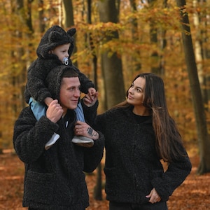 Puede incluir: Una pareja en un entorno de bosque, vistiendo chaquetas peludas negras. El hombre lleva a un ni&ntilde;o peque&ntilde;o sobre sus hombros. El ni&ntilde;o lleva una chaqueta con capucha peluda negra con orejas de oso. La pareja est&aacute; de pie sobre un lecho de hojas de oto&ntilde;o ca&iacute;das.