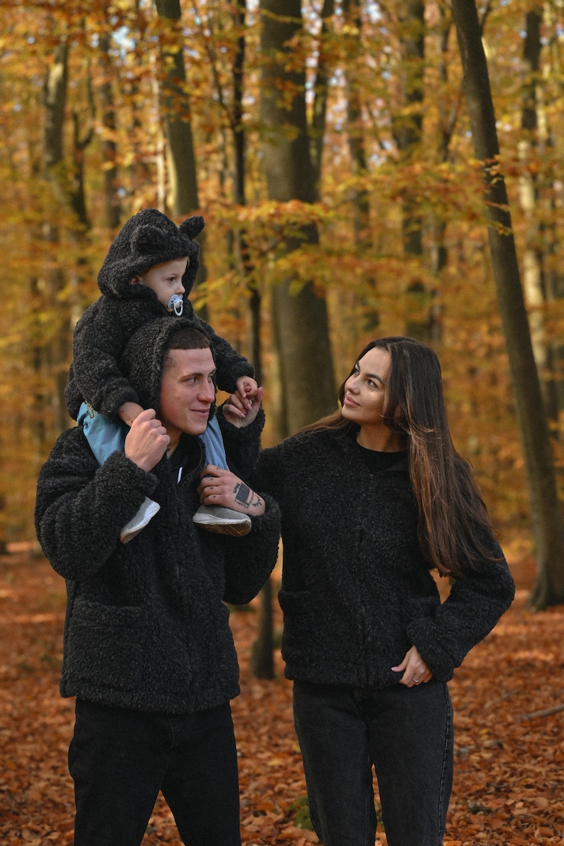 Puede incluir: Una pareja en un entorno de bosque, vistiendo chaquetas peludas negras. El hombre lleva a un ni&ntilde;o peque&ntilde;o sobre sus hombros. El ni&ntilde;o lleva una chaqueta con capucha peluda negra con orejas de oso. La pareja est&aacute; de pie sobre un lecho de hojas de oto&ntilde;o ca&iacute;das.