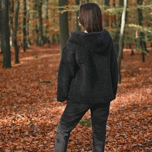 Puede incluir: Una persona con una chaqueta de vell&oacute;n negra con capucha y jeans de lavado oscuro de pie en un bosque con hojas ca&iacute;das en el suelo.