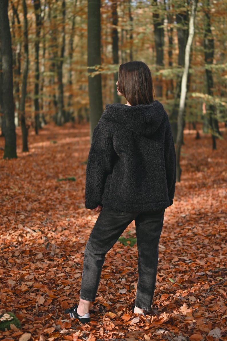 Puede incluir: Una persona con una chaqueta de vell&oacute;n negra con capucha y jeans de lavado oscuro de pie en un bosque con hojas ca&iacute;das en el suelo.