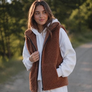 Può includere: Un gilet senza maniche in sherpa marrone con chiusura a zip nera integrale e cappuccio, indossato sopra una felpa bianca con cappuccio e maniche lunghe. Il gilet è realizzato in materiale pile testurizzato e presenta due tasche anteriori.