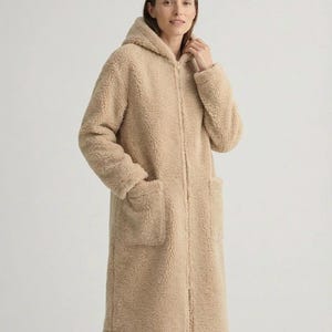 May include: A beige, knee-length, hooded coat made of a textured, fleece-like material. The coat features a full-length front zipper closure, long sleeves, and two large, square patch pockets on the front. The design is simple and functional.