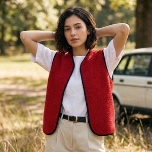 Puede incluir: Un chaleco de forro polar rojo con ribete negro se usa sobre una camiseta blanca y pantalones beige. El chaleco tiene una apariencia texturizada y un dise&ntilde;o simple y abierto. Se ve un cintur&oacute;n negro con hebilla dorada.