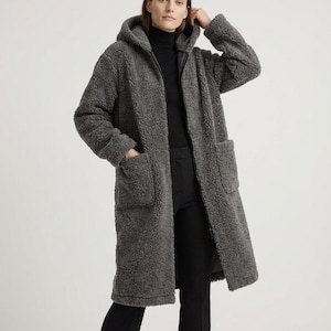 May include: A charcoal gray, knee-length coat made of a textured, fleece-like material. This long-sleeved garment features a hood, a full-length front zipper closure, and two large, square patch pockets positioned on the front of the coat.