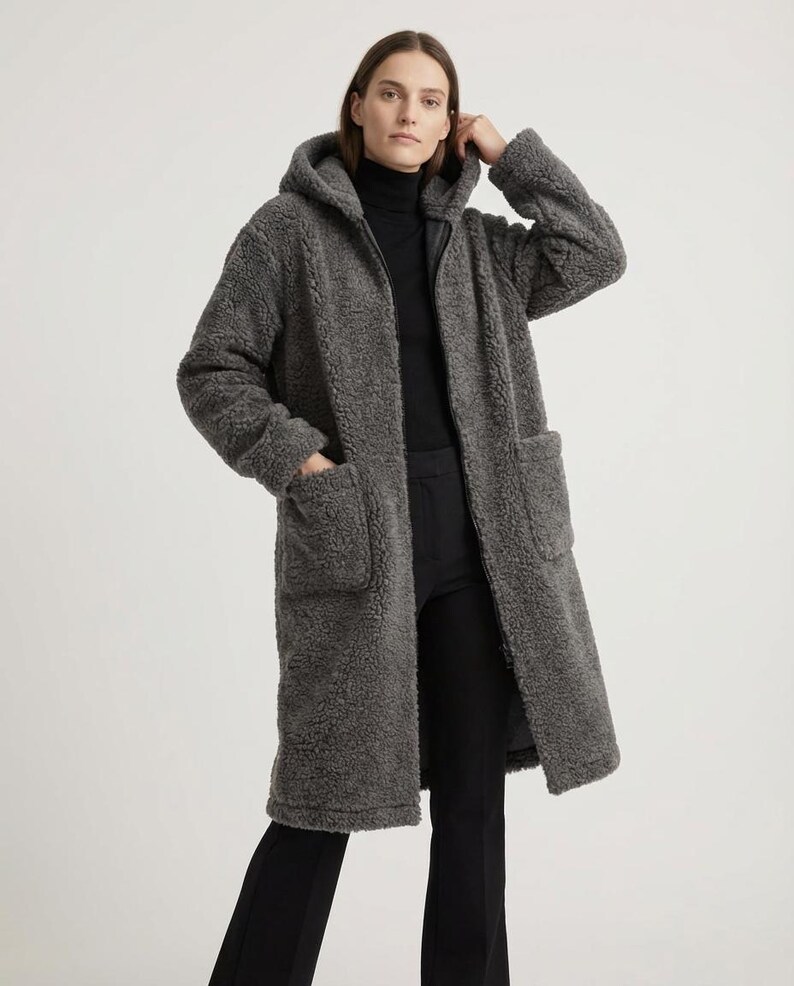 May include: A charcoal gray, knee-length coat made of a textured, fleece-like material. This long-sleeved garment features a hood, a full-length front zipper closure, and two large, square patch pockets positioned on the front of the coat.