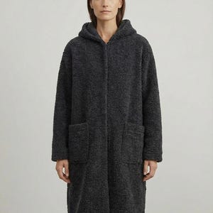May include: A charcoal gray long-length coat made of a textured, curly fleece material. The coat features a hood, a front zipper closure, and two large patch pockets on the front. The garment has long sleeves and a straight, relaxed fit silhouette.