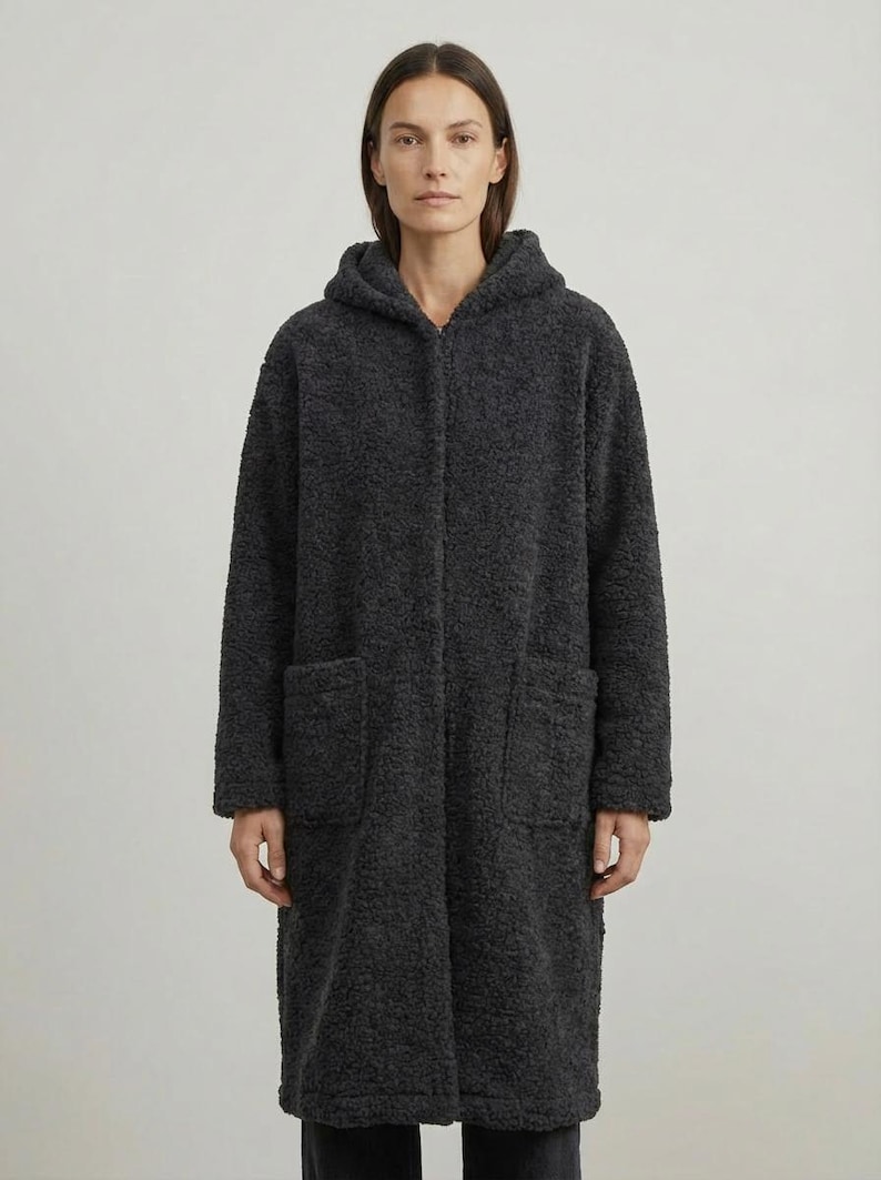 May include: A charcoal gray long-length coat made of a textured, curly fleece material. The coat features a hood, a front zipper closure, and two large patch pockets on the front. The garment has long sleeves and a straight, relaxed fit silhouette.