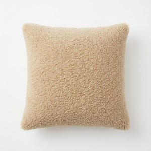May include: A square, beige-colored pillow with a textured, plush surface. The pillow has a soft, fuzzy appearance, suggesting a comfortable and cozy design. The pillow is likely used for decorative or functional purposes.