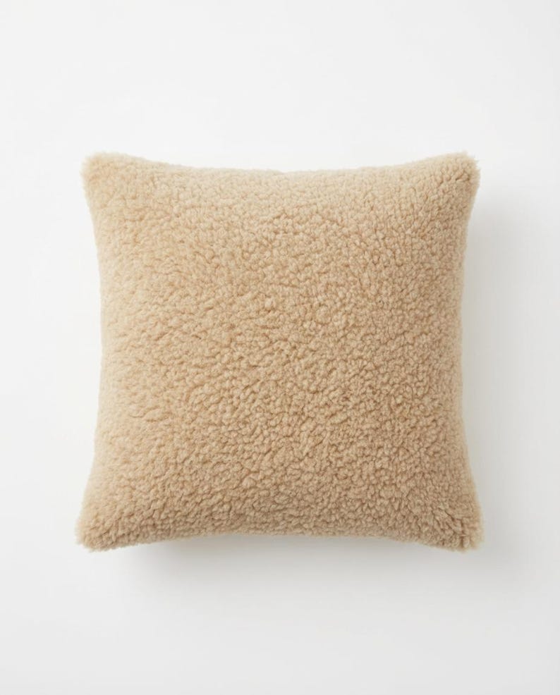 May include: A square, beige-colored pillow with a textured, plush surface. The pillow has a soft, fuzzy appearance, suggesting a comfortable and cozy design. The pillow is likely used for decorative or functional purposes.