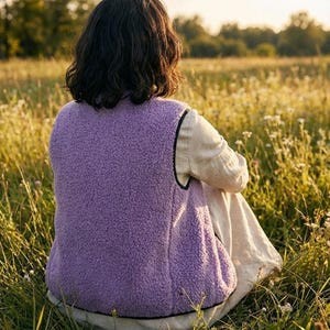 K&ouml;nnte beinhalten: Eine Person tr&auml;gt eine lavendelfarbene Fleeceweste mit schwarzem Besatz, die in einem Feld mit hohem Gras und Wildblumen sitzt. Die Weste mit weicher Textur steht im Mittelpunkt. Der Hintergrund zeigt eine verschwommene Landschaft unter warmem Sonnenlicht.