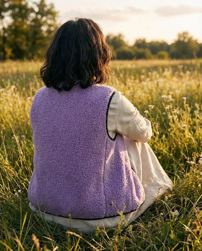 K&ouml;nnte beinhalten: Eine Person tr&auml;gt eine lavendelfarbene Fleeceweste mit schwarzem Besatz, die in einem Feld mit hohem Gras und Wildblumen sitzt. Die Weste mit weicher Textur steht im Mittelpunkt. Der Hintergrund zeigt eine verschwommene Landschaft unter warmem Sonnenlicht.