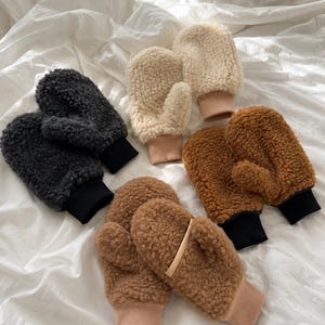 Puede incluir: Una colección de cinco pares de manoplas de forro polar sherpa difuso en gris carbón, crema y marrón. Cada par presenta un exterior grueso y texturizado con un puño de punto acanalado de color sólido en negro o beige, diseñado para el abrigo.
