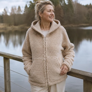 Op de afbeelding: Een beige, langemouwen vest met capuchon en ritssluiting, gemaakt van een textuurrijk, fleece-achtig materiaal. Het vest heeft een volledige ritssluiting aan de voorzijde, een capuchon en twee voorzakken. Het wordt gedragen over een licht shirt.