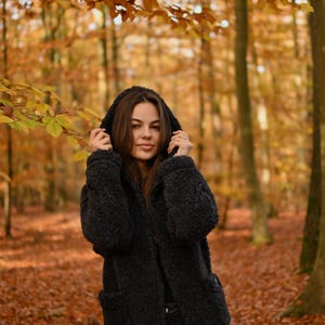 Puede incluir: Una mujer con una chaqueta negra con capucha se encuentra en un bosque con follaje oto&ntilde;al. La chaqueta tiene una textura peluda y la mujer mira a la c&aacute;mara.