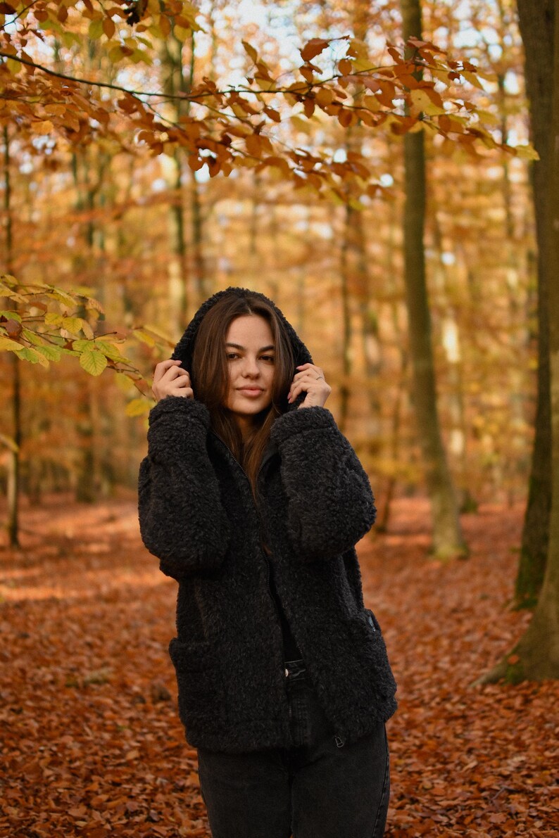 Puede incluir: Una mujer con una chaqueta negra con capucha se encuentra en un bosque con follaje oto&ntilde;al. La chaqueta tiene una textura peluda y la mujer mira a la c&aacute;mara.
