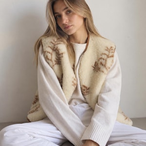 Könnte beinhalten: Eine ärmellose Weste aus cremefarbenem Schaffell mit braunem Farnmuster, getragen über einem cremefarbenen, langärmeligen Strickpullover und einer weißen Hose. Die Weste ist an den Vorderkanten und am Ausschnitt mit braunem Leder eingefasst.
