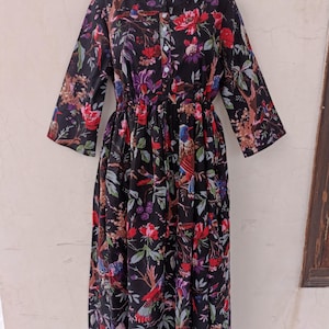 Puede incluir: Un vestido negro con estampado floral que presenta un patrón vibrante de flores rojas, moradas y azules, junto con pájaros y follaje. El vestido tiene una botonadura frontal, una cintura ceñida y mangas tres cuartos. El vestido está diseñado para un ajuste cómodo.