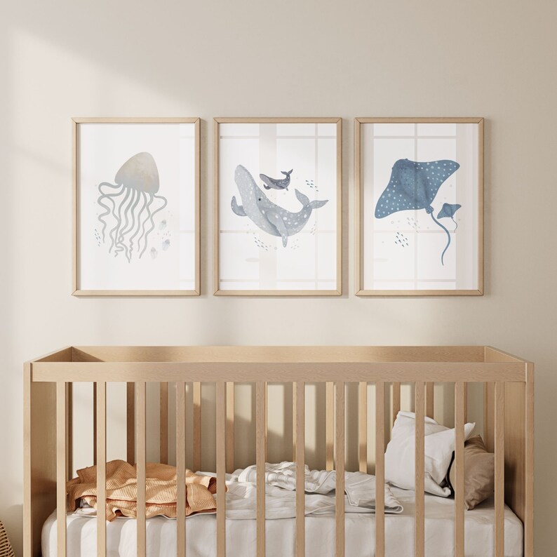 May include: Three framed watercolor prints of sea creatures. The prints feature a jellyfish, a whale, and a stingray. The prints are in a light blue and gray color scheme and are hung on a white wall above a wooden crib.