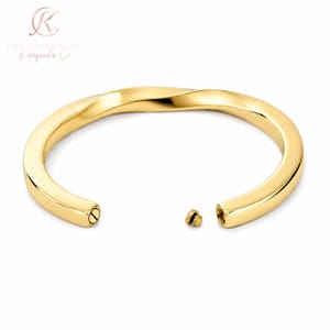 Könnte beinhalten: Ein goldfarbener Armreif aus Metall mit einem glatten, gedrehten Design. Das Armband verfügt über einen hohlen Innenraum, der durch eine kleine, mit einem Gewinde versehene Schraubkappe zugänglich ist, die abgenommen zwischen den Enden liegt.