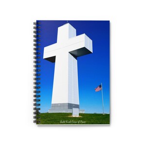 Può includere: Una grande croce bianca si erge contro un cielo blu brillante. La croce è un monumento noto come Bald Knob Cross of Peace. Una piccola bandiera americana sventola su un palo in primo piano.