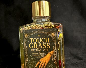 May include: A clear glass bottle with a gold dropper cap containing yellow oil and dried botanical pieces. The black rectangular label features gold text reading TOUCH GRASS RITUAL OIL, STRESS RELIEF, ANXIETY HELP, and WITCH ELIXIR, with an illustration of a hand touching grass.