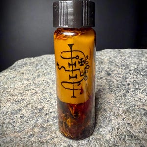 May include: A small clear glass vial with a black screw-top lid, filled with a thick yellow substance and dark botanical matter at the base. A black sigil-like symbol is printed on the front of the yellow layer within the glass container.