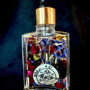 May include: A clear glass bottle with a gold cap and black dropper, filled with colourful flower petals and a white charm. The bottle contains red, blue, and yellow petals. The charm has a black design.