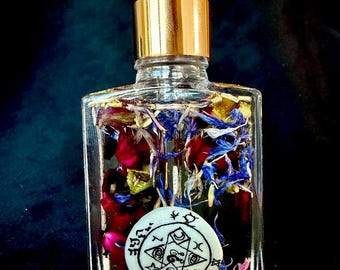 May include: A clear glass bottle with a gold cap and black dropper, filled with colorful flower petals and a white charm. The bottle's contents include red, blue, and yellow petals. The charm has a black design.