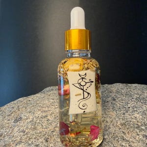 Könnte beinhalten: Eine klare Glasflasche mit goldenem Verschluss und weißer Pipette, gefüllt mit einer Flüssigkeit und getrockneten Blumen. Ein weißes Etikett mit schwarzer Schrift ist an der Flasche befestigt. Die Flasche steht auf einer grauen Steinoberfläche.