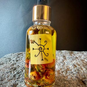 May include: A clear glass dropper bottle filled with yellow oil, dried orange flower buds, and green herbs. A rectangular label is attached to the front, featuring a black hand-drawn spiral symbol with radiating lines. The bottle has a gold collar.