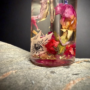Könnte beinhalten: Eine klare Glasflasche gefüllt mit getrockneten Blumen und Kräutern. Die Flasche enthält rote, rosa und gelbe Blütenblätter sowie einen weißen Papierauschnitt mit einem Augendesign. Die Flasche steht auf einer grauen Steinoberfläche.
