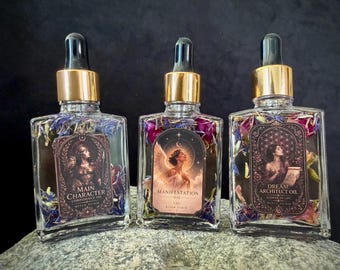 Op de afbeelding: Drie vierkante glazen flessen met gouden druppeldopjes, gevuld met heldere olie en gedroogde bloemblaadjes. De etiketten bevatten gedetailleerde illustraties en de tekst: Main Character, Manifestation en Dream Architect Oil.