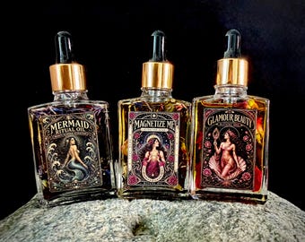 Op de afbeelding: Drie vierkante glazen druppelflesjes gevuld met olie en gedroogde botanische elementen. De etiketten bevatten illustraties van een zeemeermin, een vrouw met een ster en een vrouw met een spiegel, gelabeld als Mermaid Ritual Oil, Magnetize Me en Glamour Beauty Ritual Oil.