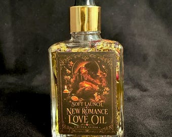 May include: A clear glass dropper bottle filled with yellow oil and dried flower petals. The label features an illustration of a person and the text: SOFT LAUNCH NEW ROMANCE LOVE OIL WITCH ELIXIR. The bottle has a gold collar and a black dropper top.