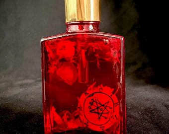 Op de afbeelding: Een rechthoekige glazen fles gevuld met een felrode vloeistof en zwevende botanische elementen. De fles heeft een goudkleurige hals en een zwarte druppelaar. Een rond rood etiket met een zwart occult symbool is op de voorzijde bevestigd.