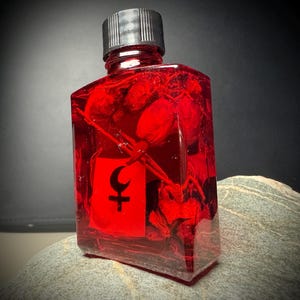 Puede incluir: Una botella rectangular transparente llena de líquido rojo y bayas rojas. La botella tiene una tapa negra y una etiqueta roja con un símbolo negro de luna creciente y cruz. La botella está sobre una piedra gris.