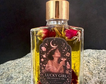 Op de afbeelding: Een doorzichtige glazen fles met een gouden druppeldop, gevuld met gele olie en gedroogde rode rozenknoppen. Het etiket toont een illustratie van een persoon met een glas, een halve maan en de tekst LUCKY GIRL ABUNDANCE OIL WITCH ELIXIR.