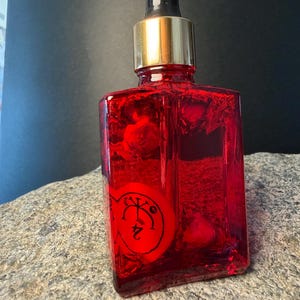 Peut inclure: Une bouteille en verre rouge avec un compte-gouttes noir et un bouchon doré. La bouteille contient un liquide rouge et un objet circulaire rouge avec des symboles noirs et le chiffre 24. La bouteille est posée sur une surface en pierre grise.
