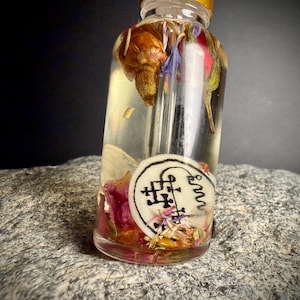 Op de afbeelding: Een heldere glazen fles gevuld met een lichtgekleurde vloeistof en gedroogde bloemen, waaronder rode rozenblaadjes en blauwe korenbloemen. Een wit etiket met zwarte symbolen is op de fles bevestigd. De fles staat op een grijs, getextureerd stenen oppervlak.