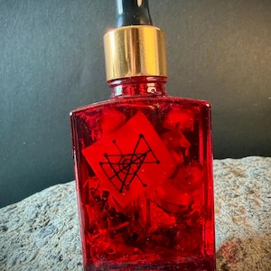 Könnte beinhalten: Eine rechteckige rote Glasflasche mit goldenem Verschluss und schwarzer Pipette. Die Flasche enthält rote Flüssigkeit und ein kleines rotes Papier mit einem schwarzen geometrischen Design. Die Flasche steht auf einer Steinoberfläche.