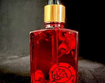 Op de afbeelding: Een vierkante glazen fles gevuld met een felrode vloeistof en zwevende botanische elementen. De fles heeft een goudkleurige hals, een zwarte druppelaar en een rond rood etiket met een zwart lijntekening-zegel en de letters N en P.