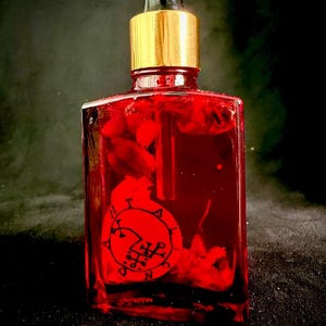 Op de afbeelding: Een rechthoekige glazen fles gevuld met een felrode vloeistof en zwevende botanische elementen. De fles heeft een goudkleurige hals en een zwarte druppelaar. Een rond rood etiket op de voorzijde toont zwarte tekst en een zegelontwerp.